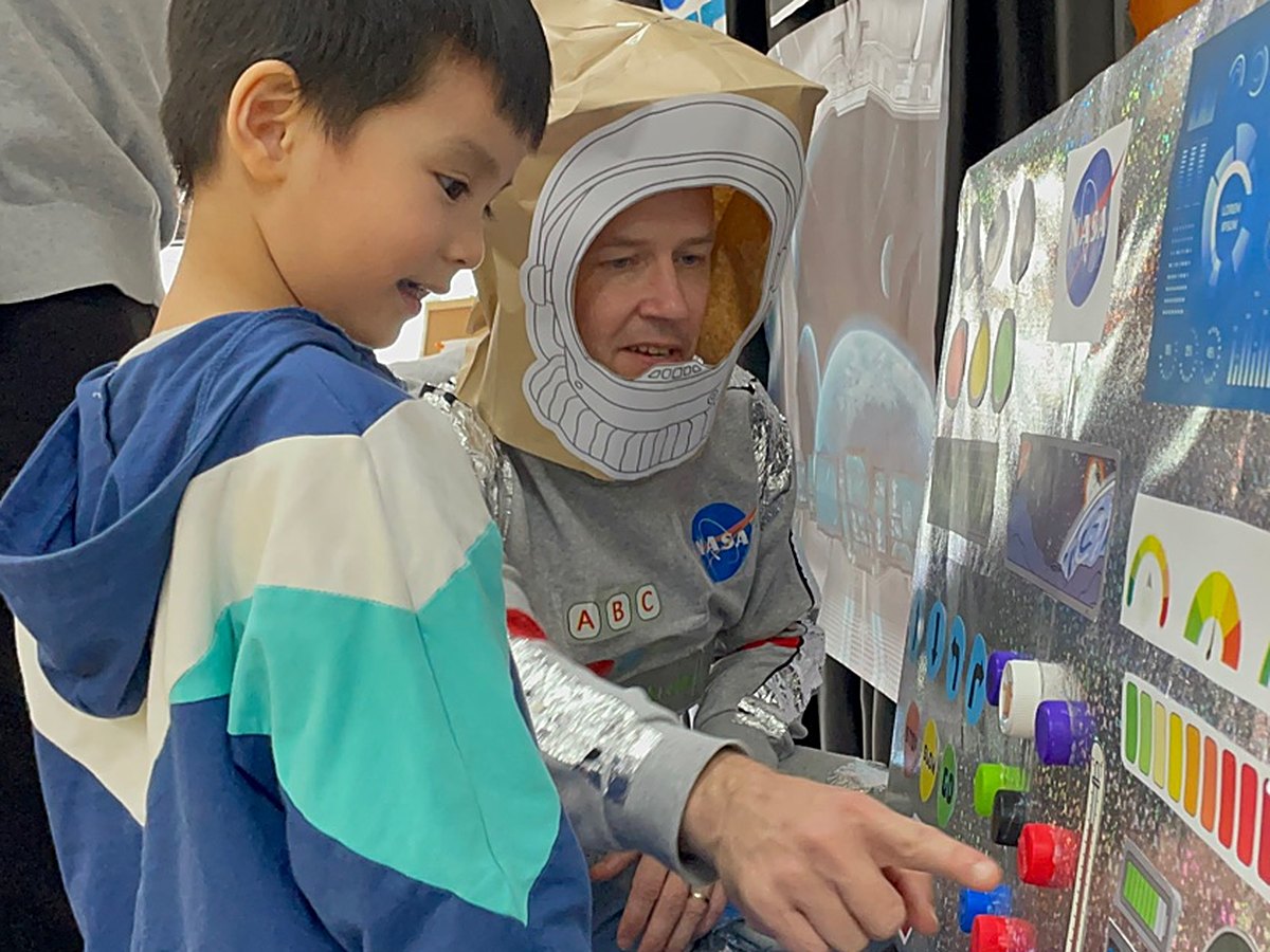 A child pressing buttons on a space control panel with his teacher dressed as an astronaut